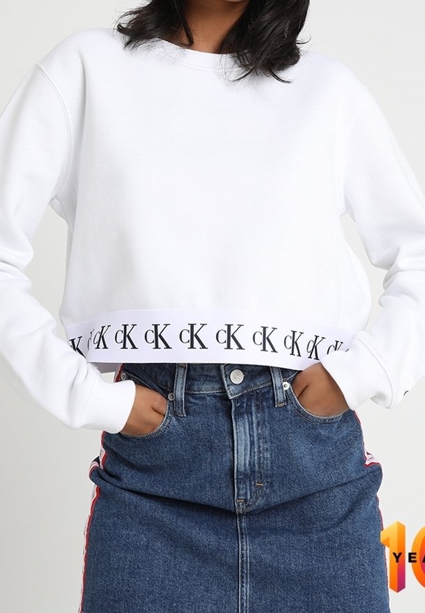 Personne portant un sweat-shirt court blanc avec le logo noir "cK" répété sur l'ourlet et une jupe en denim bleue taille haute avec des bandes rouges sur les côtés.