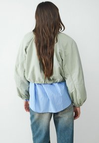 Jasnozielona kurtka bomberka z elastycznymi mankietami, nałożona na niebieską koszulę w paski, w połączeniu z wyblakłymi niebieskimi dżinsami, prezentująca swobodny styl.