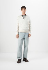 Junger Mann steht, trägt einen cremefarbenen Halbzip-Pullover über einem grauen Shirt, hellblaue Jeans und dunkelbraune Schnürschuhe vor einem weißen Hintergrund.