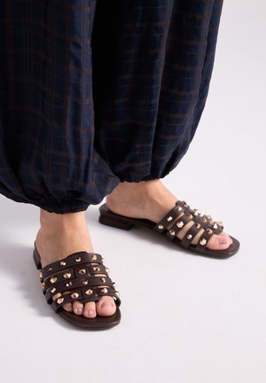 Pies con sandalias marrón oscuro con tachuelas doradas, combinadas con pantalones sueltos de cuadros azul marino recogidos en los tobillos.