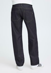 Jeans in denim blu scuro con vestibilità dritta, caratterizzati da cuciture gialle e design a cinque tasche, indossati con sneakers bianche.