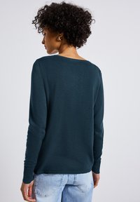 Dunkelgrüner Pullover mit langen Ärmeln, weitem Ausschnitt und gerippten Bündchen. Über hellblauen Jeans getragen, die eine glatte Textur zeigen.