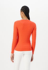 Pull torsadé rouge avec des poignets et un ourlet côtelés ; présente une coupe ajustée et des manches longues. Le modèle porte un pantalon blanc, de dos par rapport à l'appareil photo.