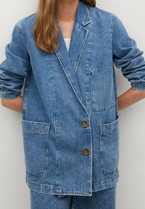 Frau trägt ein locker sitzendes, blaues Denim-Blazer mit zwei Fronttaschen und braunen Knöpfen, kombiniert mit passenden Denim-Hosen und einem weißen Unterhemd.