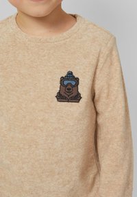 Kind trägt einen beigen Pullover mit gesticktem Bären, der eine blaue Wintermütze und eine Schutzbrille auf der Brust trägt.