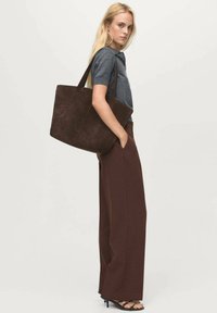 Bolso tote de gamuza marrón con asas largas, combinado con pantalones de pierna ancha marrones oscuros y una camiseta de manga corta gris. Textura suave, diseño minimalista.