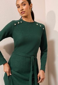 Zelené žebrované šaty s dlouhými rukávy, páskem v pase a dekorativními knoflíky na ramenou. Textura je měkká a design je přiléhavý.