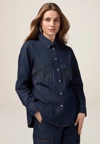 Camicia di denim blu scuro con colletto a bottone, caratterizzata da un design verticale di perline scintillanti sul fronte, maniche lunghe e una vestibilità rilassata.