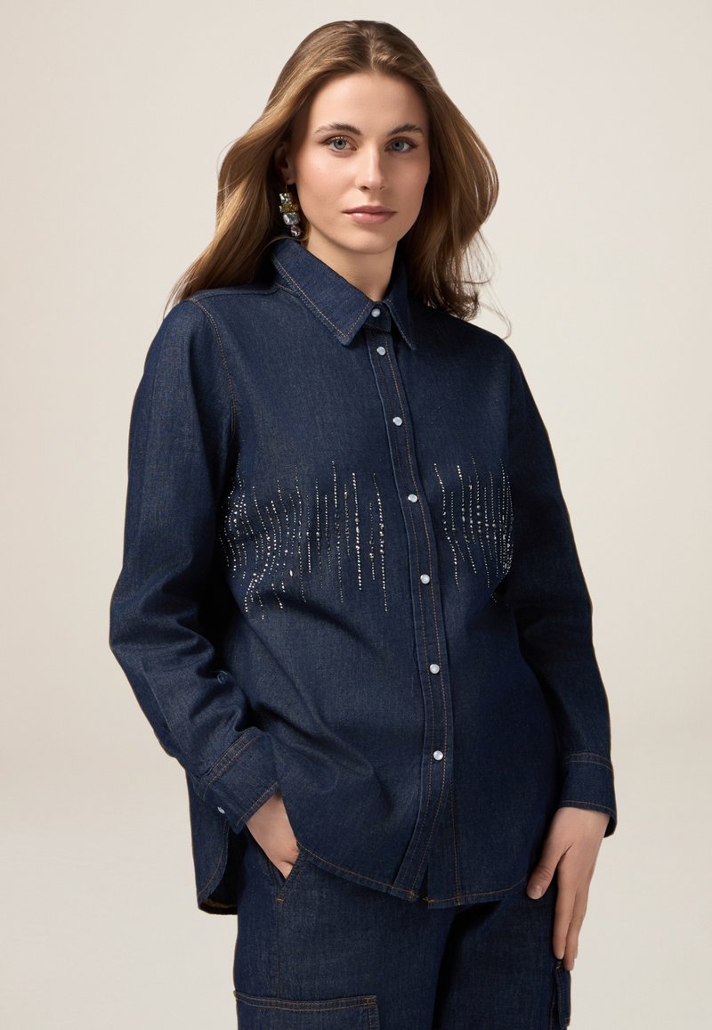 Cămașă din denim de culoare albastru închis, cu guler cu nasturi, având un design strălucitor cu mărgele verticale pe partea din față, mâneci lungi și o croială lejeră.