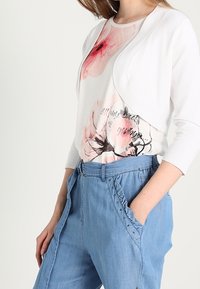 Chaqueta corta blanca sobre una camisa blanca con estampado floral en tonos rosas y detalles en negro, combinada con unos pantalones de mezclilla azul claro, holgados.