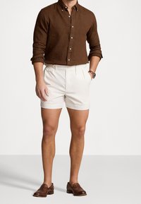 Uomo che indossa una camicia marrone a maniche lunghe con bottoni, pantaloni corti a vita alta color panna, mocassini in pelle marrone e un orologio da polso, in piedi con una mano in tasca.