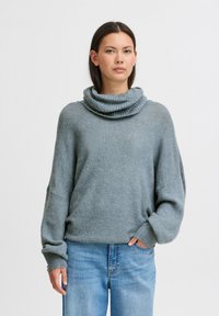 Maglione oversize grigio chiaro con collo a scialle, polsini e orlo a costine; realizzato in un morbido tessuto testurizzato; abbinato a jeans azzurri chiari.
