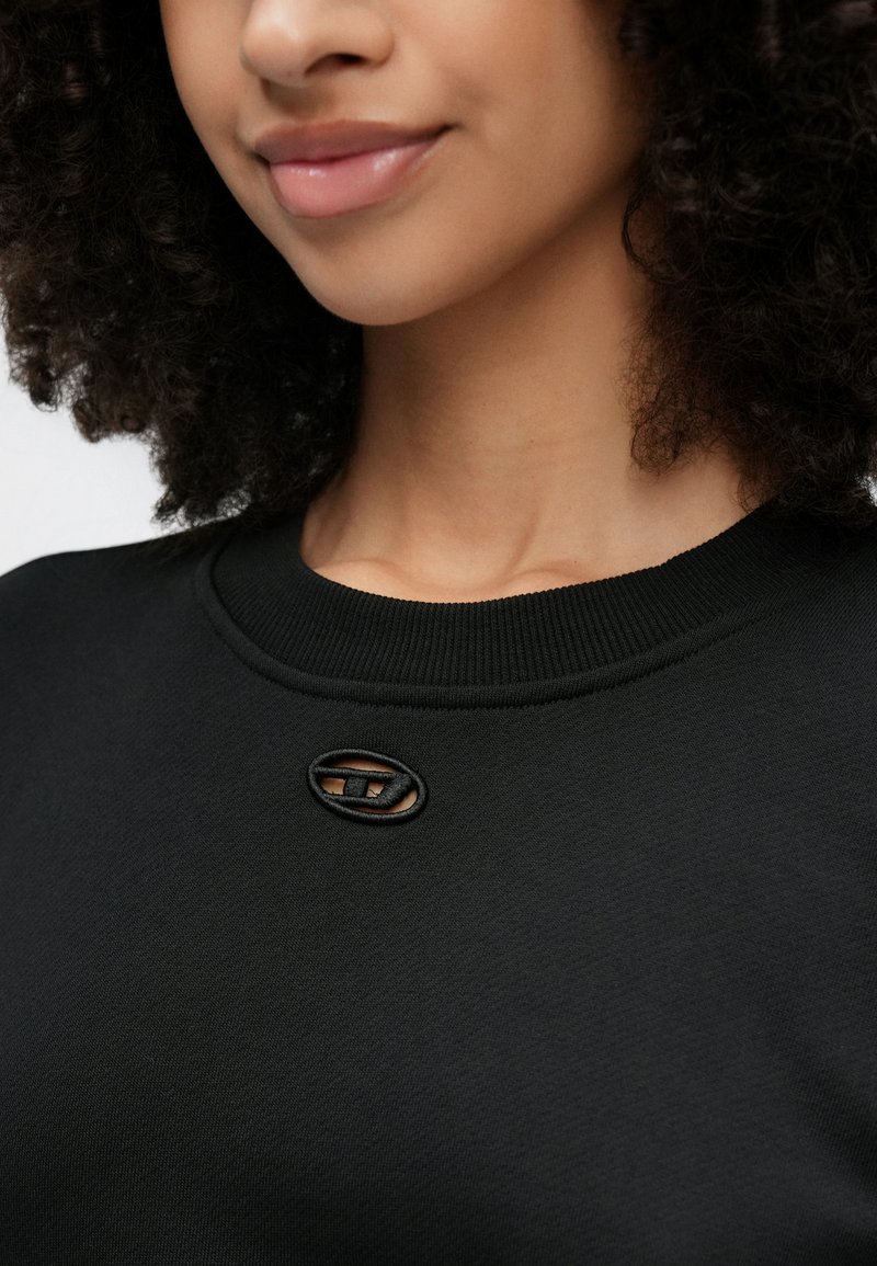 Donna con capelli ricci che indossa una felpa nera con un piccolo taglio ovale decorativo vicino allo scollo.