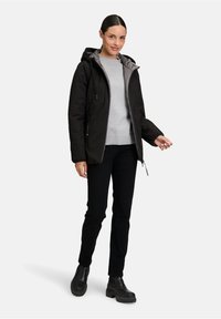Veste noire imperméable avec une doublure intérieure grise, fermeture éclair à l'avant, capuche réglable et poches latérales, associée à un pull gris et un pantalon noir.