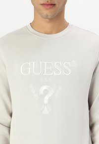 Sudadera de color gris claro con el logo bordado de "GUESS", un signo de interrogación y acentos geométricos. Textura suave y diseño de cuello redondo.