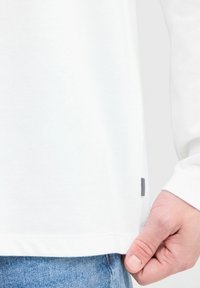Camiseta blanca de manga larga confeccionada en suave algodón, con un dobladillo redondeado y una pequeña etiqueta en blanco y negro en un lado. Mano visible.