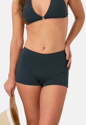 Kobieta w ciemnoszarych kąpielówkach i dopasowanym górnym bikini, trzyma beżowy kapelusz przeciwsłoneczny z białą opaską.