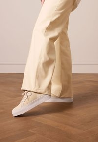 Beige sneakers met een mesh-textuur, een witte rubberen zool en veters, gecombineerd met lichtgele wijde broek, geplaatst op een houten vloer.