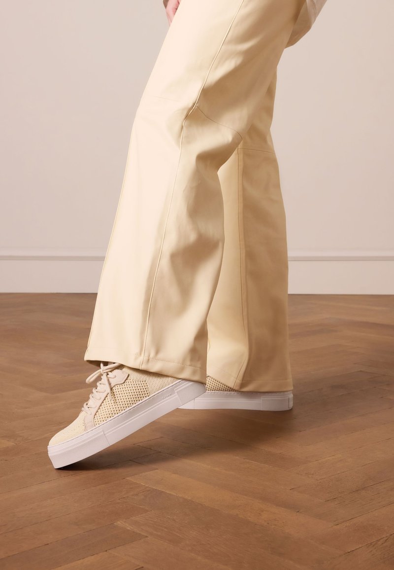 Beige sneakers met een mesh-textuur, een witte rubberen zool en veters, gecombineerd met lichtgele wijde broek, geplaatst op een houten vloer.