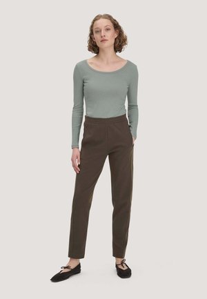 Femme aux cheveux bouclés portant un haut vert clair à manches longues, un pantalon marron taille haute et des ballerines noires, debout avec une main dans la poche.
