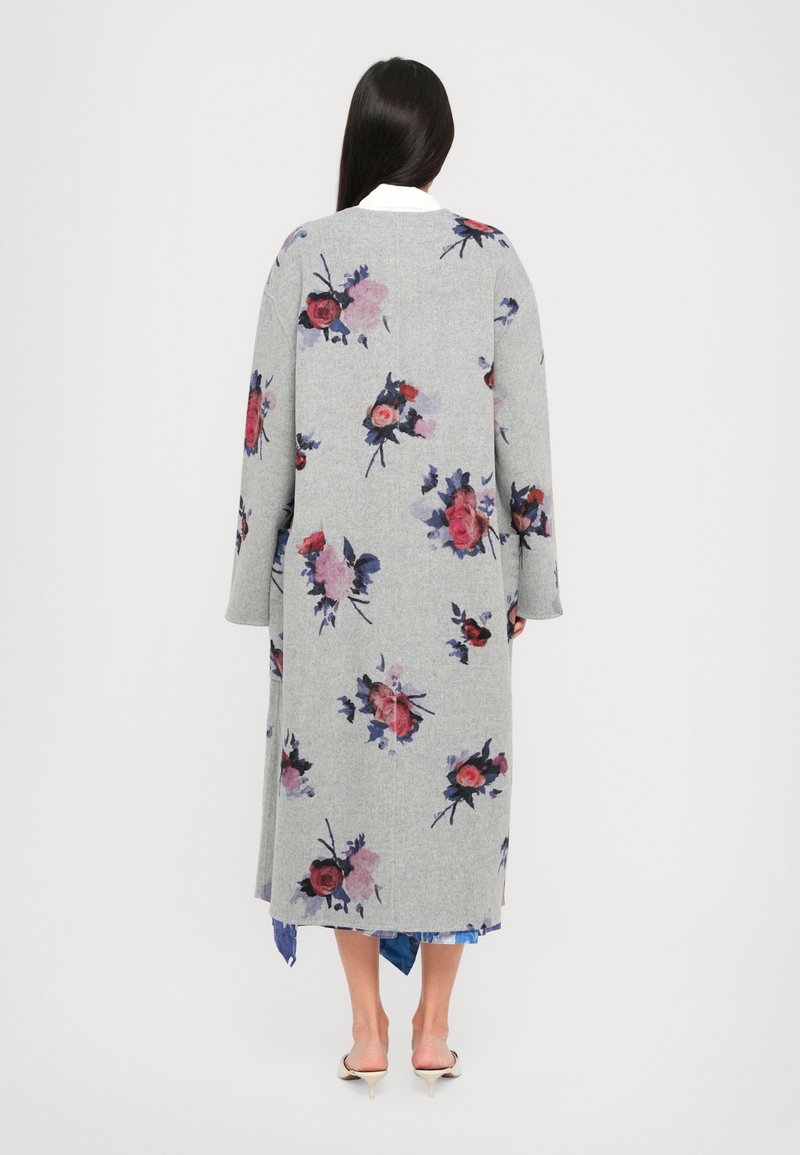 Donna con lunghe chiome scure che indossa un lungo cappotto floreale grigio, una camicia bianca, una gonna blu e scarpe con tacco kitten bianche, in piedi di spalle.