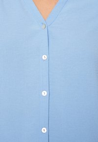 Camicia azzurra chiaro realizzata in morbido tessuto, con scollatura a V e una fila di bottoni bianchi lungo la parte frontale. Tessuto liscio, design casual.