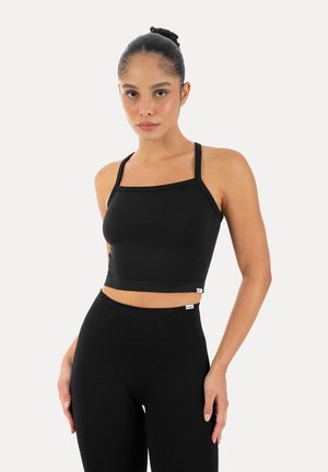Frau trägt ein schwarzes, bauchfreies Tanktop mit dünnen Trägern und passende hochgeschnittene Leggings, steht vor einem schlichten hellen Hintergrund.