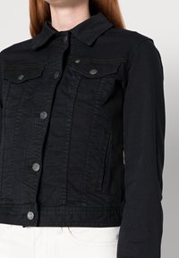 Black denim jacket featuring a button-up front, two chest pockets, and snap buttons. The design is cropped with a classic collar and textured fabric.