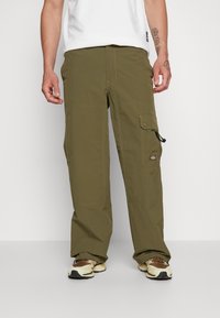 Man som bär olivgröna cargobyxor med sidofickor, vit t-shirt och bruna samt beiga sneakers, stående framför en vit bakgrund.