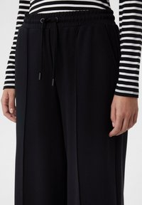 Pantalon de survêtement noir avec une taille élastique et un cordon de serrage, doté de poches latérales et d'une texture lisse. Haut à manches longues rayé visible.
