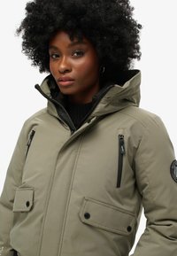 Oliwkowa kurtka ocieplana z kapturem, z kieszeniami na zamek błyskawiczny i kieszeniami na klapkę, czarnymi zamkami błyskawicznymi oraz okrągłą naszywką z logo na rękawie. Gładka tekstura.