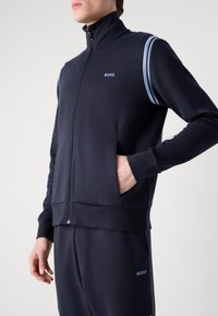 Jūras spēku zips aizverams sporta jakas izgatavota no gludas auduma, ar zilu akcents svītrām uz pleciem un logo uz krūšu daļas, ar priekšējiem kabatām.