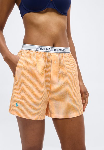 Γυναίκα που φοράει πορτοκαλί και άσπρο ριγέ μπόξερ σορτς με ζώνη μέσης Polo Ralph Lauren και σκούρο μπλε σουτιέν, με τα χέρια στις τσέπες.