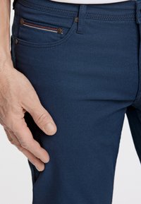 Marineblaue strukturierte Hose mit einer Fronttasche, die einen rot-weiß-blauen Streifen zeigt. Sichtbare Knopf- und Nähdetails. Hand auf der Tasche platziert.