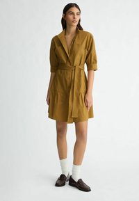 Robe chemise couleur moutarde avec une taille cintrée, un devant à boutons et des manches courtes bouffantes. Elle présente une jupe évasée et des poches latérales.