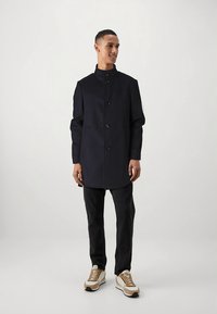 Sötétkék kabát magas gallérral, elöl gombos zárral és oldalzsebekkel, sötét farmerrel és krémszínű tornacipővel párosítva. Simaságot sugárzó anyag, letisztult dizájn.