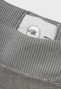 Grå sweatshirtkrage med ribbad struktur, utrustad med en sydd vit etikett och en genomskinlig knapp ovanför halsringningen. Mjuk tygdetalj synlig.