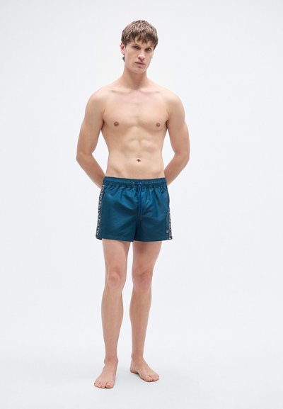 Ελαστικό σορτς κολύμβησης σε τιρκουάζ με μπλε κορδόνι μέσης και διακοσμητικές πλευρές με μοτίβα. Το μοντέλο στέκεται ξυπόλητο.