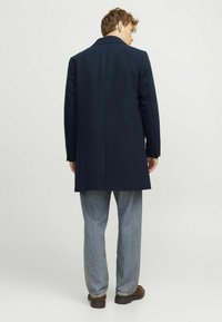 Abrigo navy con un diseño estructurado, ventilación trasera sencilla y cuello de solapa, combinado con jeans azul claro y zapatos marrones.