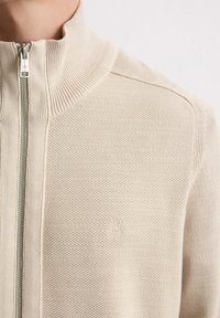 Beige ziptrui met een ribbelkraag, gestructureerde brei-stof en een klein reliëflogo op de borst. Rits is zilverkleurig.