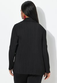 Zwarte geplooide blazer van textuurstof, lange mouwen en met een kraag. De achterkant toont een rechte snit en een gladde afwerking. Rode oorbellen zichtbaar.