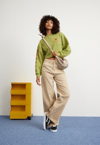 Olive green cropped sweatshirt with red graphic, beige wide-leg pants, and black sneakers. Model holds a beige shoulder bag. Yellow storage unit in background.