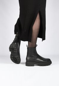 Botas de tobillo de cuero negro con suela de goma gruesa, con un tirador y paneles laterales elásticos. Llevadas con una prenda negra que muestra una abertura.