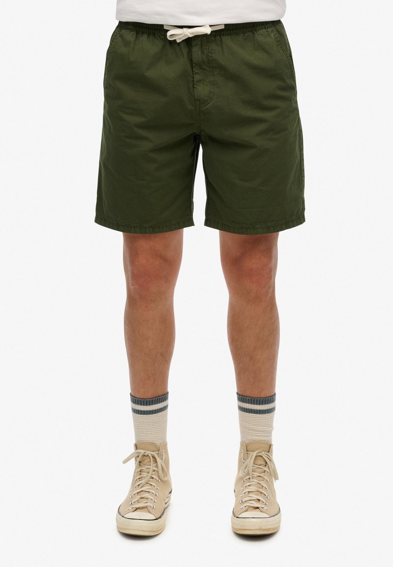 Pantalones cortos de algodón verde con cintura elástica y cordón, presentando bolsillos laterales y un corte recto. Llevados con zapatillas altas beige.
