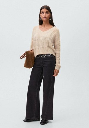 Suéter beige de punto con escote en V, combinado con unos pantalones vaqueros negros de pierna ancha de talle alto con detalles de tachuelas. Bolso de ante marrón y zapatos negros.