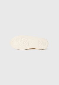 Såle av en beige gummisko med geometriske mønster og teksturerte gripeområder på en enkel lys bakgrunn.