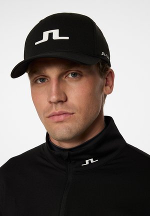 Sort kasket med et hvidt logo på fronten, der har et struktureret design og en buet skygge. Modellen bærer en sort zip-up jakke med et lille logo.