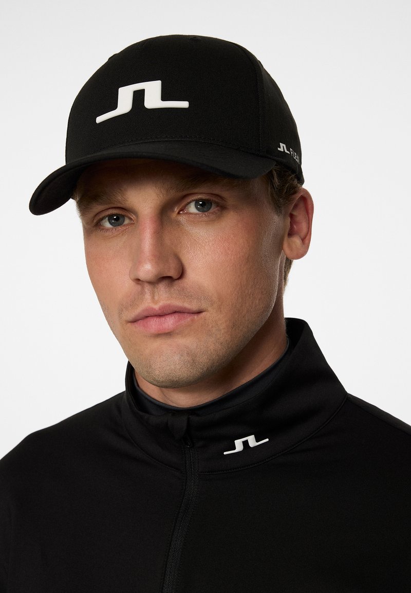 Svart keps med en vit logotyp på framsidan, med en strukturerad design och böjd skärm. Modellen bär en svart zip-up jacka med en liten logotyp.