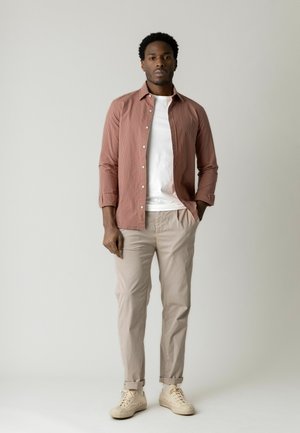 Mann steht mit den Händen in den Taschen, trägt ein staubrosa Hemd über einem weißen T-Shirt, beigefarbene Hose und weiße Turnschuhe.