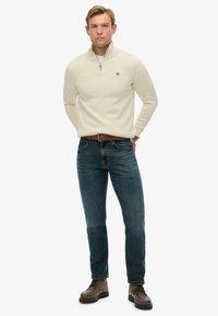 Cremefarbener Strickpullover mit hohem Kragen und Reißverschluss, kombiniert mit blauen Jeans und braunen Schnürstiefeln. Auf dem Pullover befindet sich ein kleines Logo.